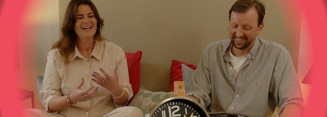 Een man en vrouw zitten lachend bovenop een bed met voor hen een wekker met het woord stress erop