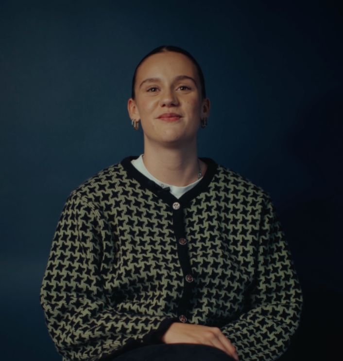 Emily zit op een stoel, met haar handen liggend op haar benen en kijkt lachend in de camera