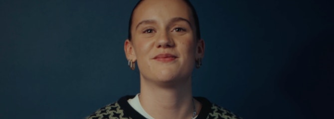 Emily zit op een stoel, met haar handen liggend op haar benen en kijkt lachend in de camera