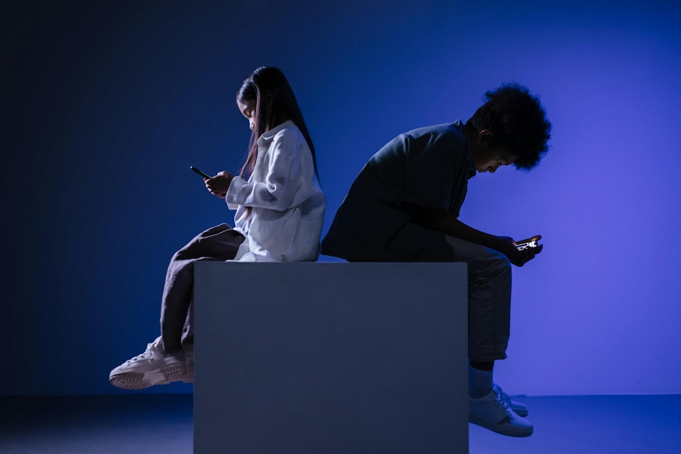 Twee jongeren zitten op een blok met hun rug tegen elkaar, beiden starend naar hun telefoon
