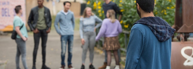 Een persoon staat alleen buiten en kijkt van afstand naar een kletsende vriendengroep