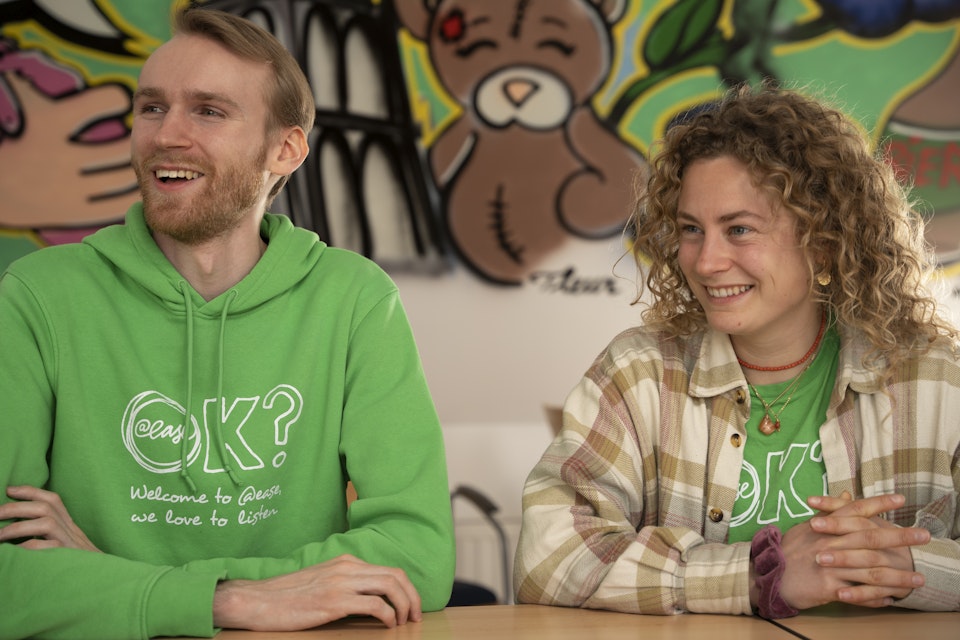Twee vrijwilligers van @ease zitten lachend naast elkaar aan een tafel