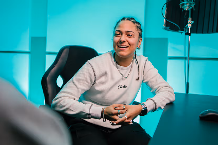 Joy zit op een bureaustoel aan een tafel en kijkt naar een persoon aan de rechterkant. Op de achtergrond is een verlichte blauwe muur.