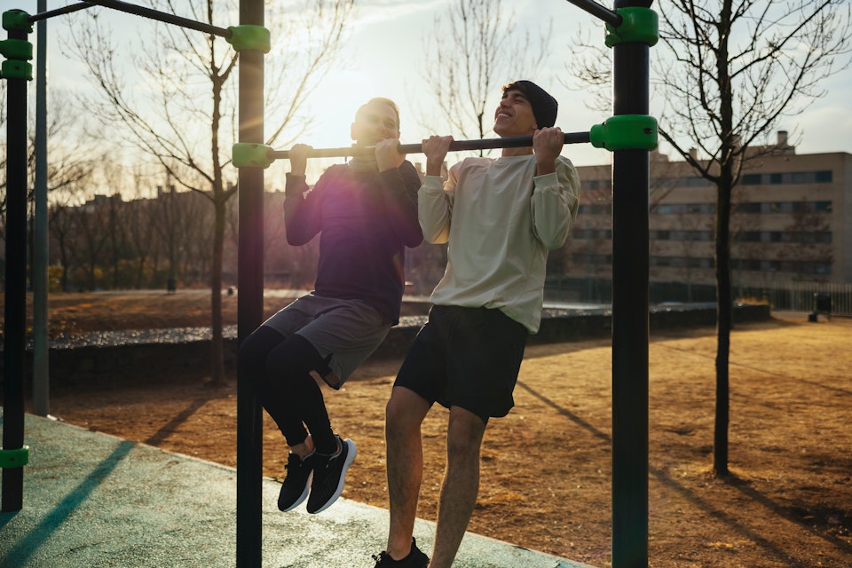 Twee personen doen buiten optrekoefeningen aan een sporttoestel in een park.