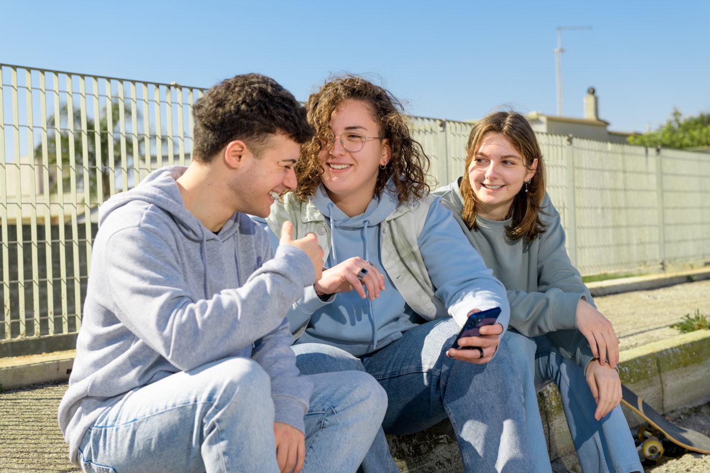 Drie lachende jongeren zitten buiten, lachen naar elkaar en hebben een telefoon vast.