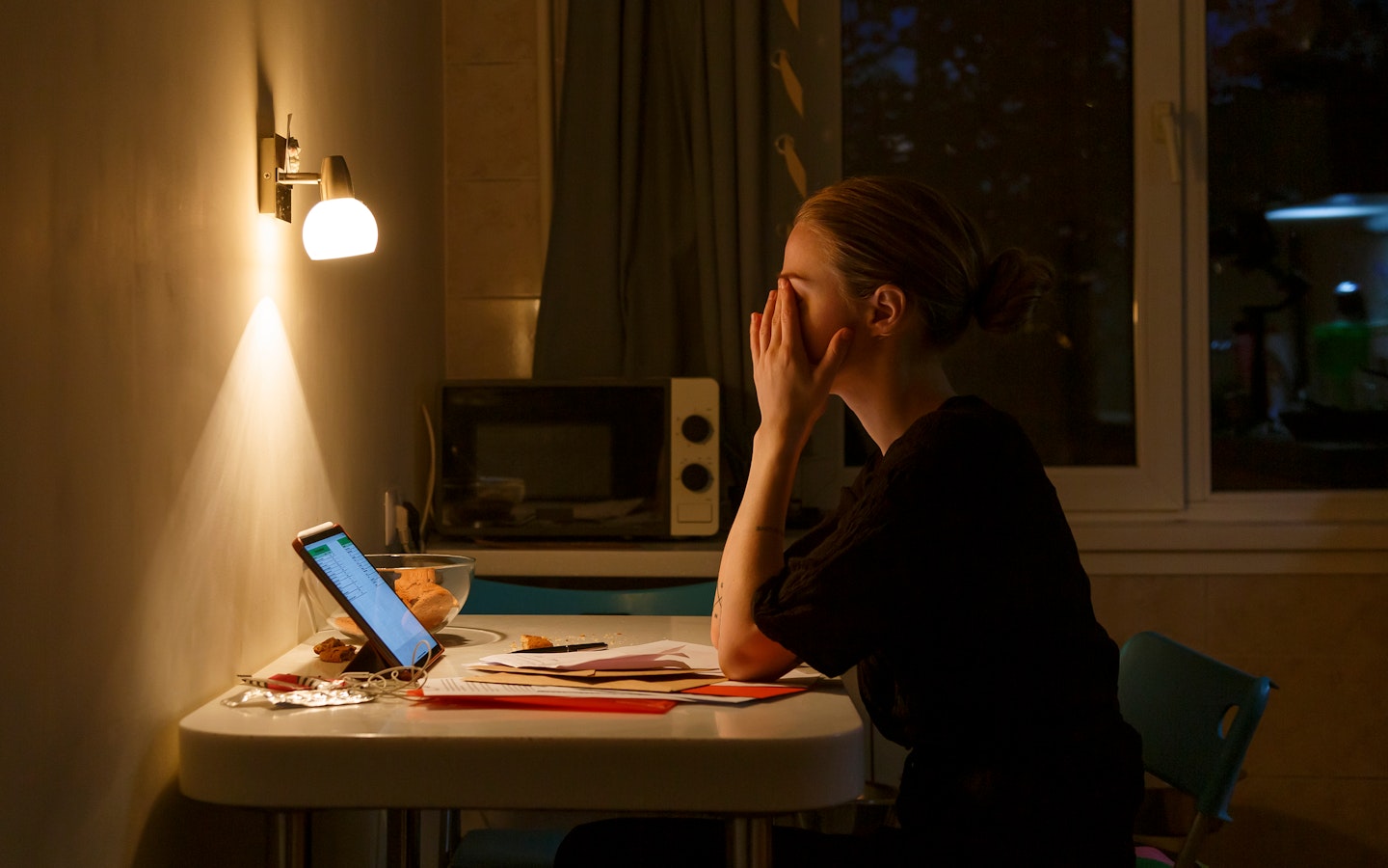Vermoeide vrouw zit in het donker achter haar laptop met haar handen voor haar gezicht.