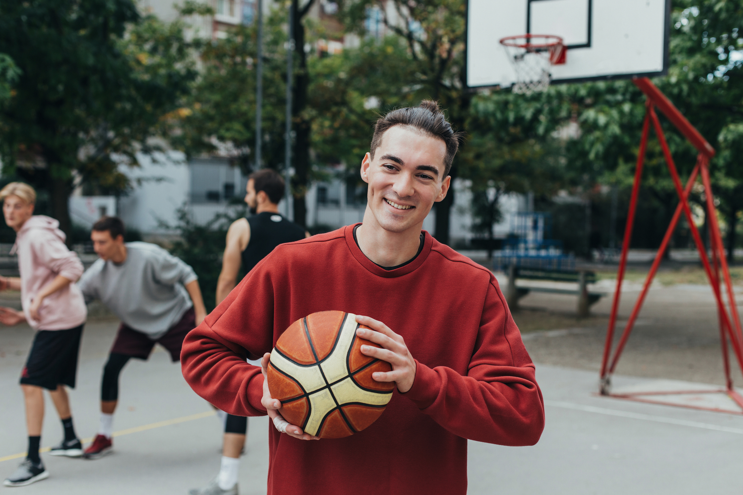 Jongeman op een buitenbasketbalveld lachend met een basketbal in zijn hand en vrienden achter hem