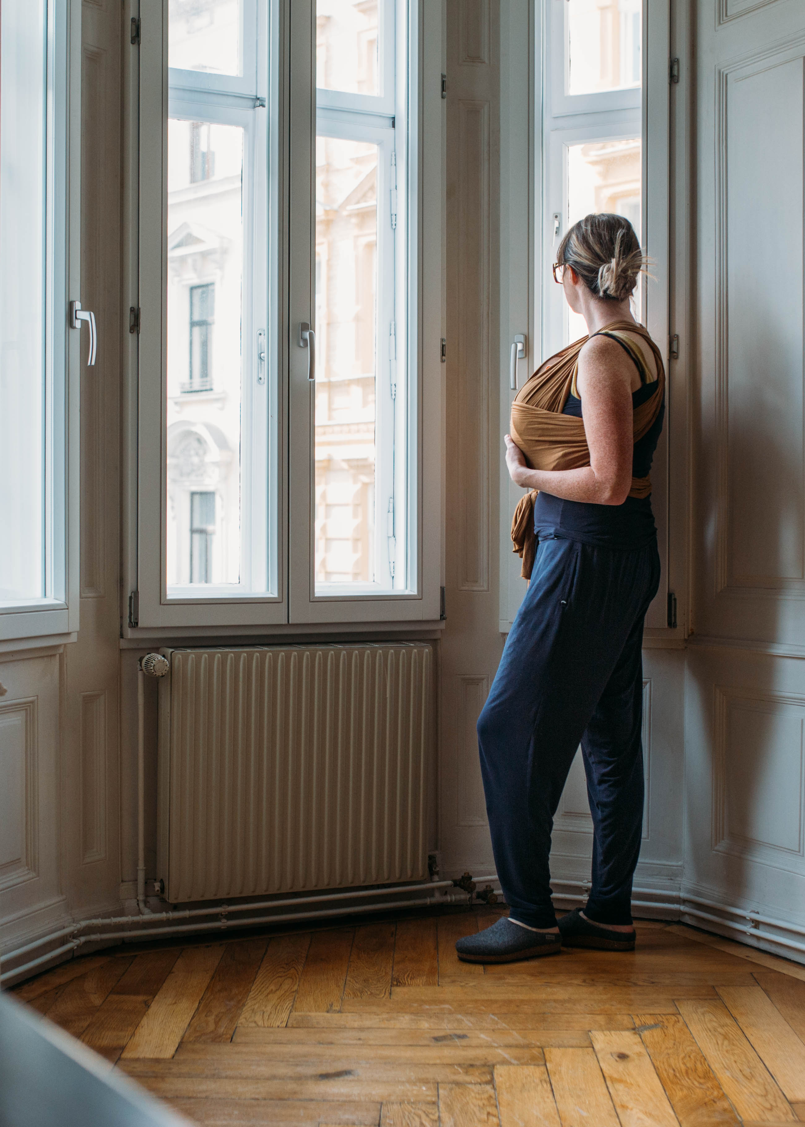 Vrouw draagt haar baby in een draagdoek en kijkt naar buiten door het raam