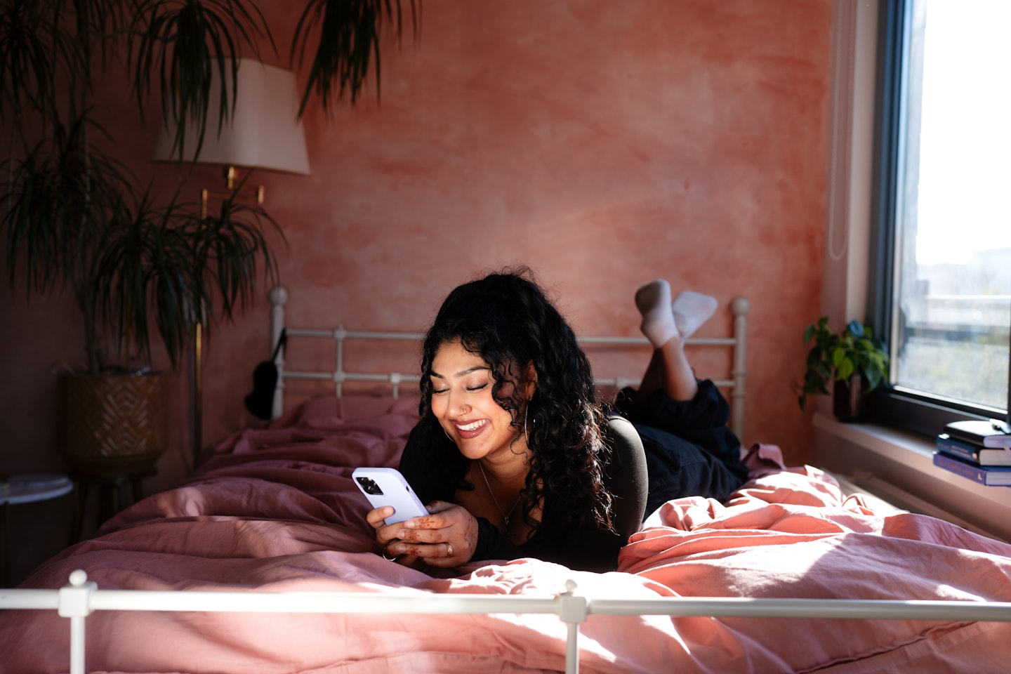 Een lachende vrouw ligt relaxed met haar telefoon op bed in een zonverlichte slaapkamer