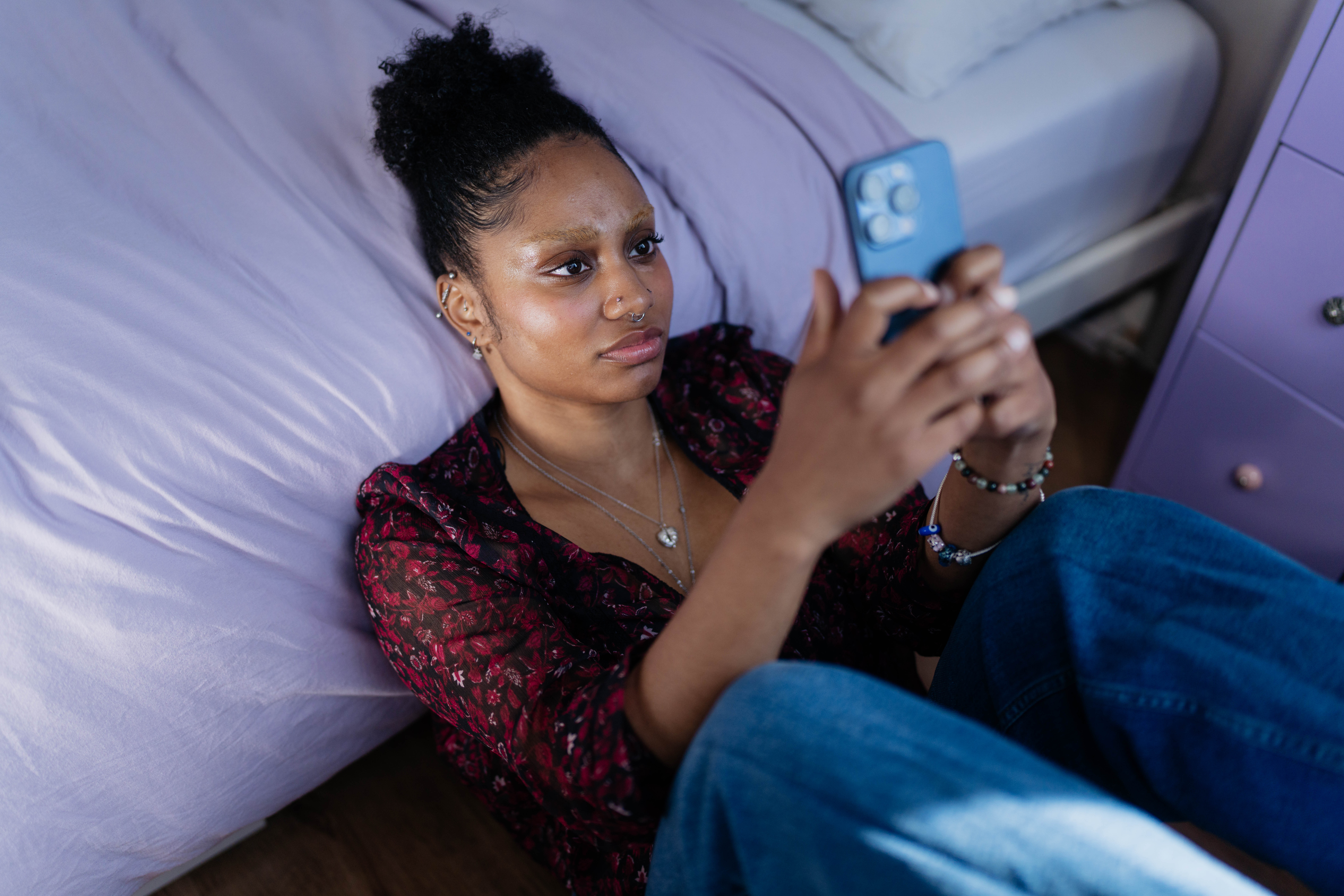 Een jonge vrouw zit rustig achterover tegen haar bed en staart naar de telefoon in haar handen