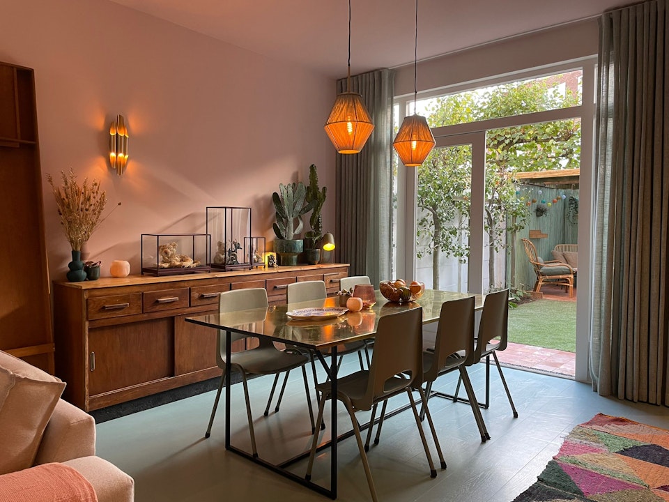 Gezellige eetkamer met glazen tafel, houten dressoir, warme hanglampen en uitzicht op de tuin.