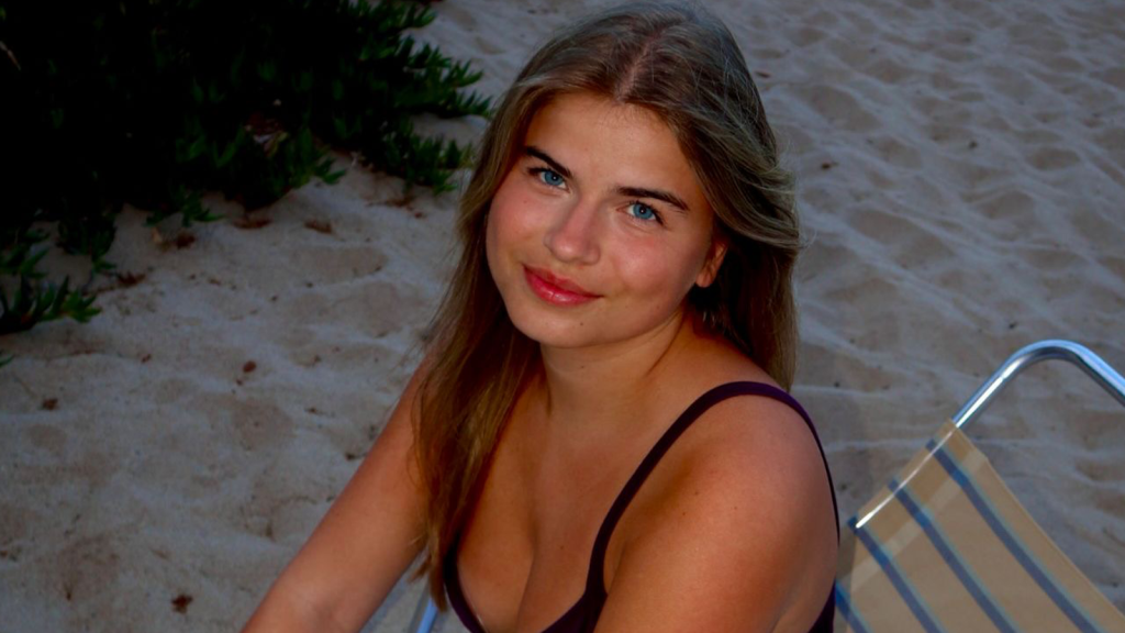 Vrouw zit op een strandbed en kijkt glimlachend in de camera.
