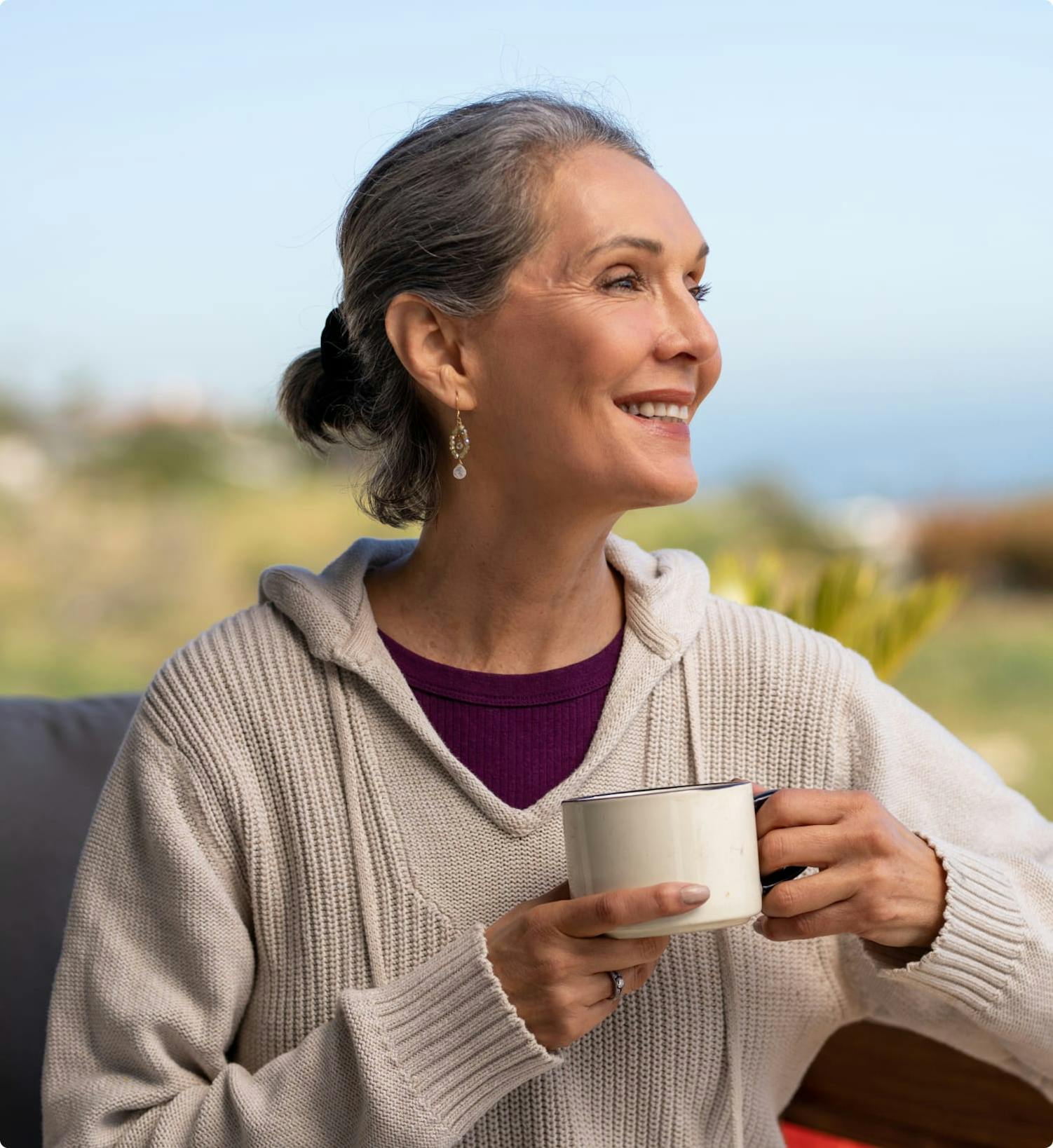 Woman sitting on a deck, holding a coffee cup, looking at the sea.