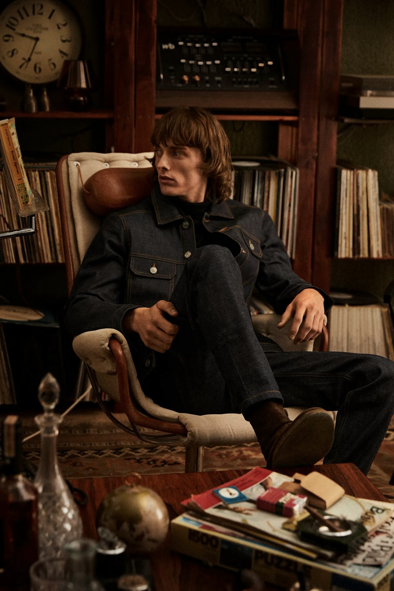 A young man wearing a denim jacket and jeans sits relaxed in a cushioned chair in a cozy, retro-styled room.