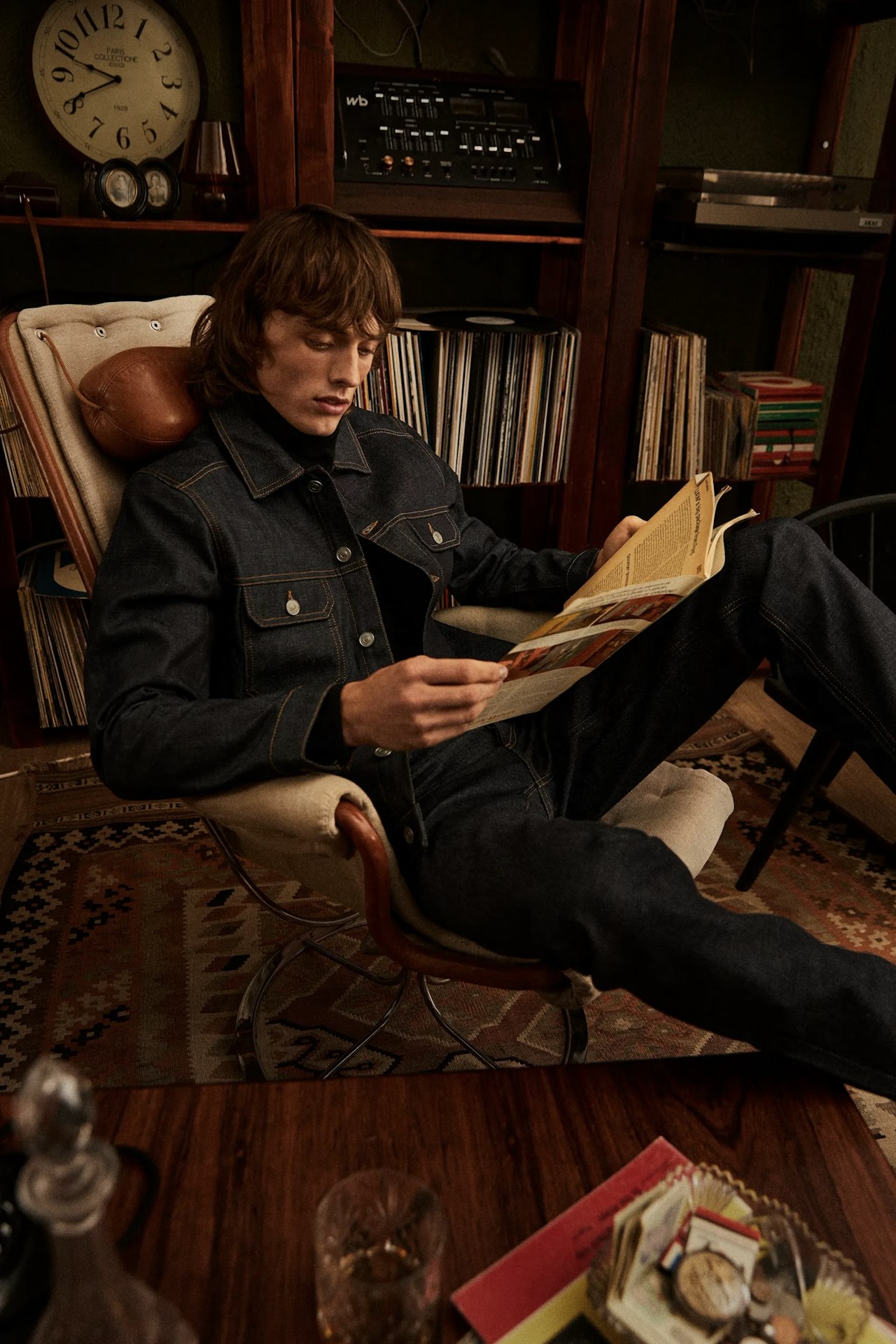A young man in a denim jacket and jeans relaxes in a lounge chair, reading a magazine