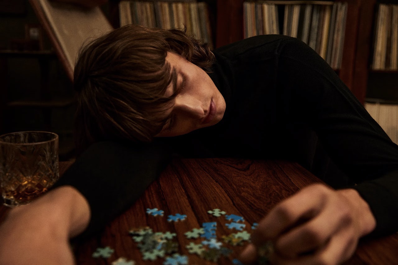A young man with medium-length brown hair in a dark turtleneck rests their head and arm on a wooden table beside a scattered jigsaw puzzle