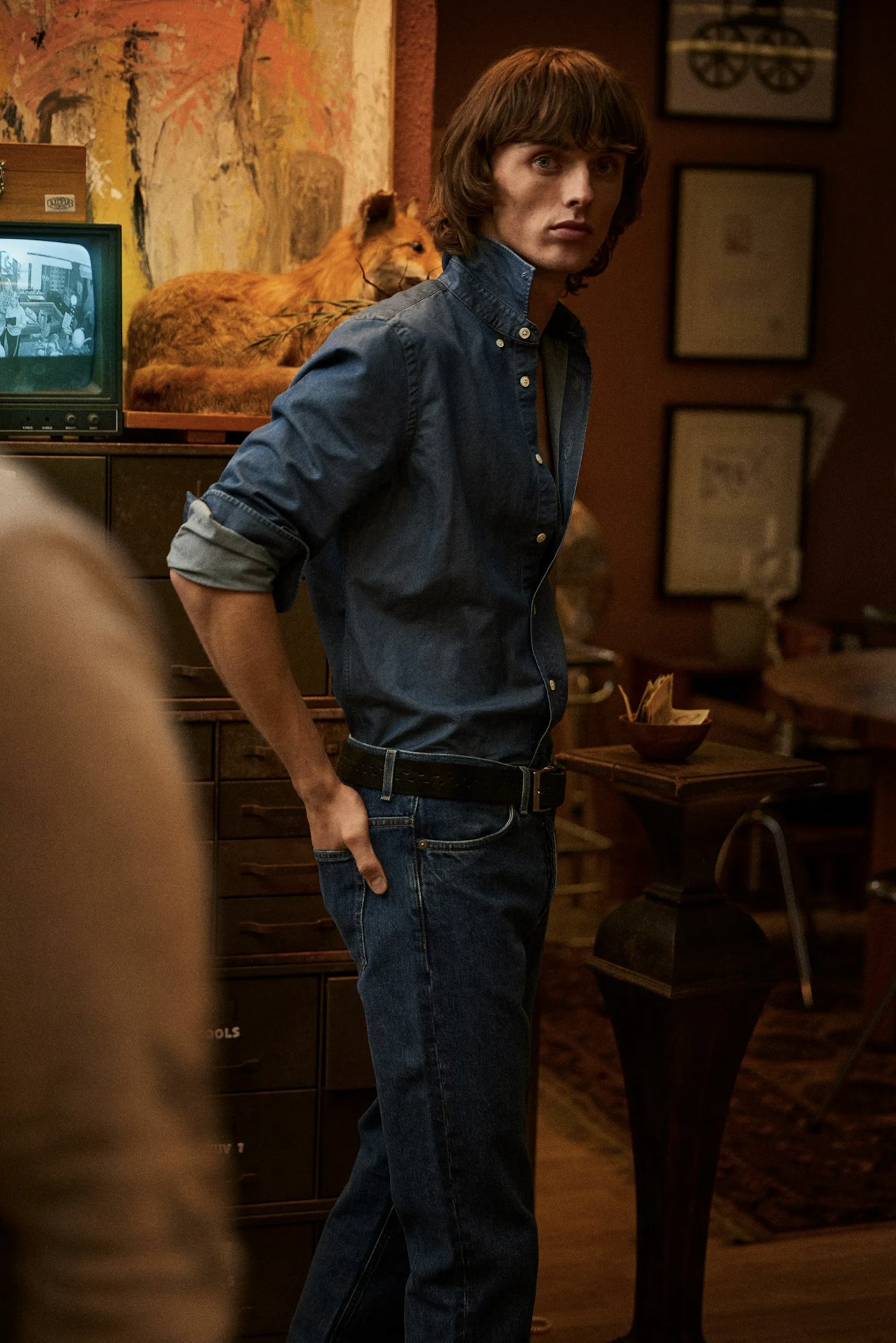 A young man with medium-length brown hair, wearing a denim shirt and jeans, stands indoors with one hand in their back pocket