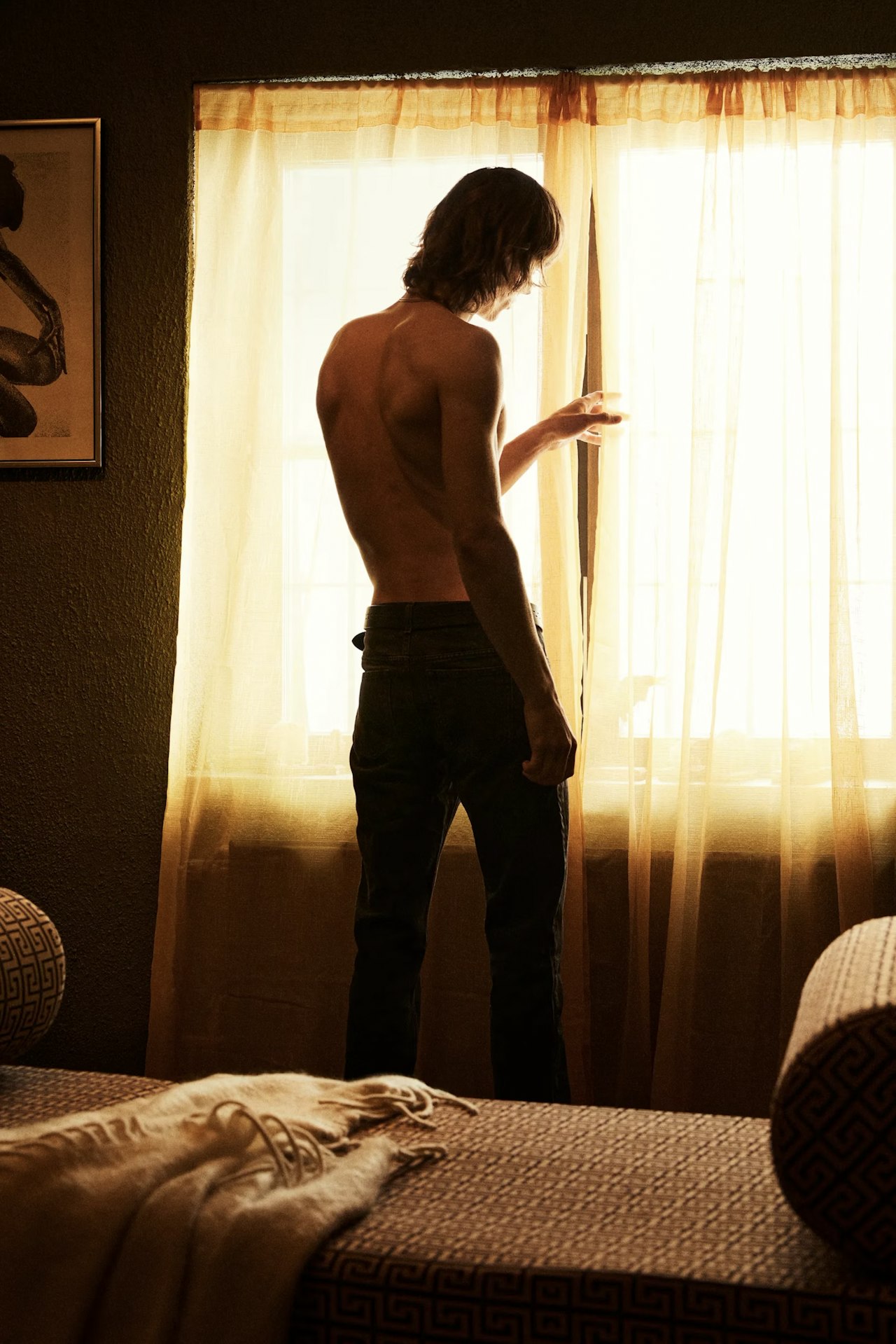 A shirtless man stands by a window with sheer curtains, looking outside into bright sunlight