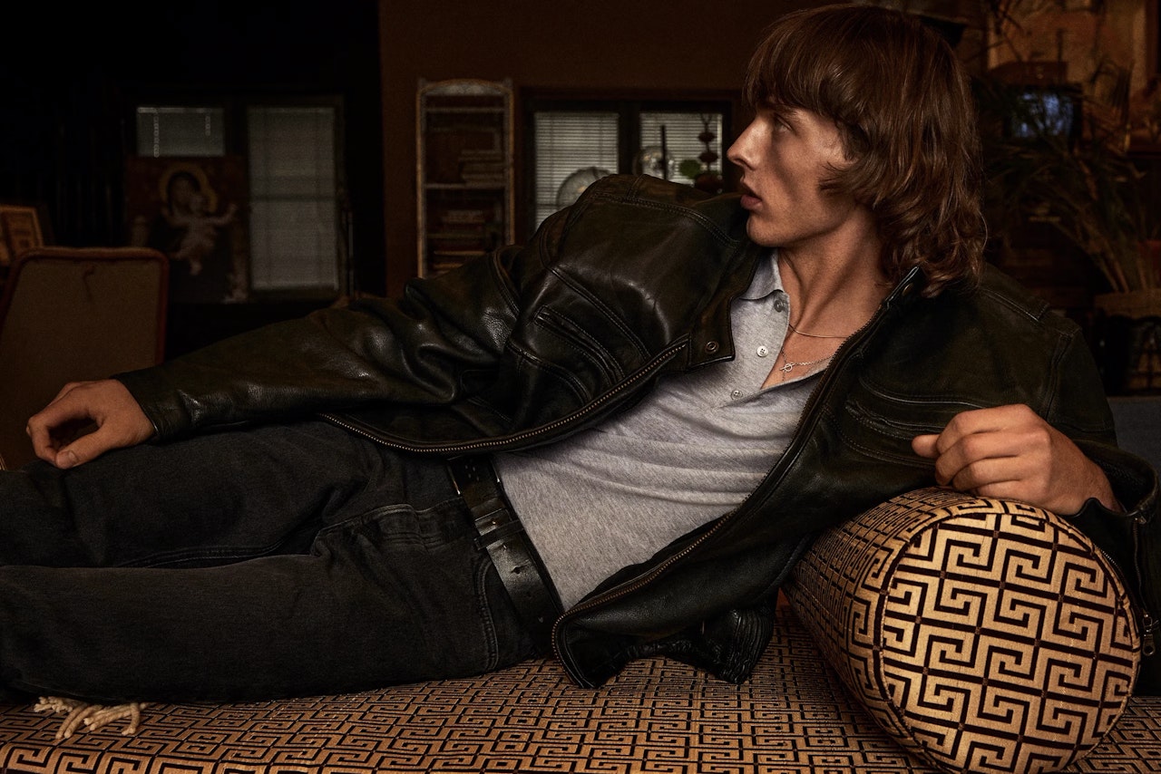 A man with brown hair, wearing a black leather jacket and gray shirt, reclines sideways on a patterned couch, looking off to the right in a dimly lit room.