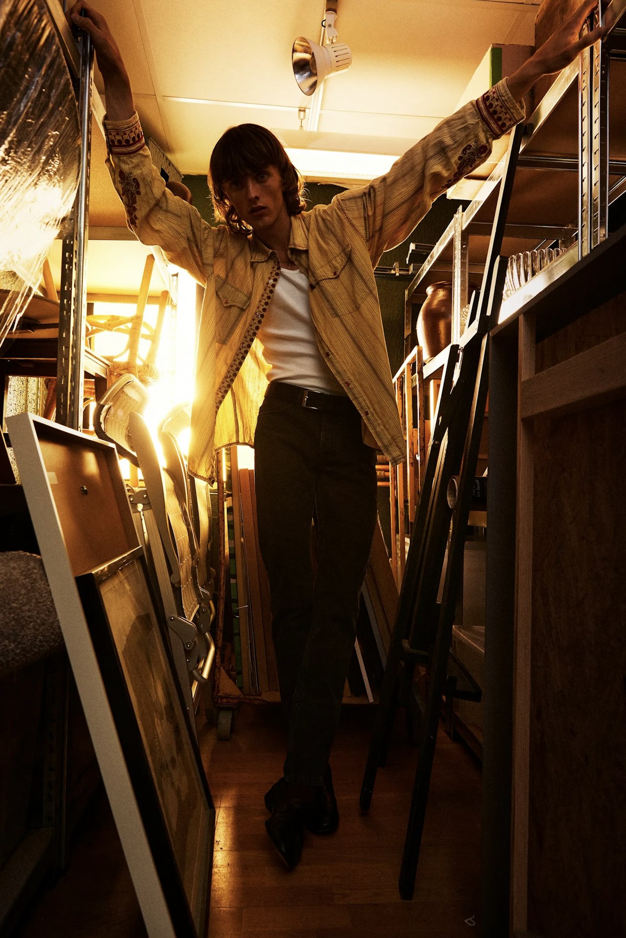 A young man in a yellow shirt leans with arms outstretched between metal shelves in a cluttered room