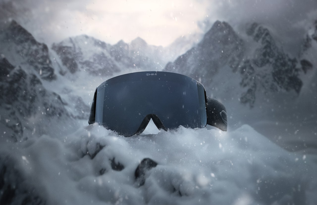 Black ski goggles resting in the snow with snowy mountains in the background.