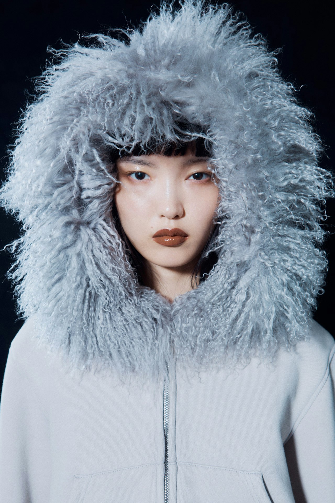 Woman in a fluffy gray hooded jacket with a neutral expression on a dark background.