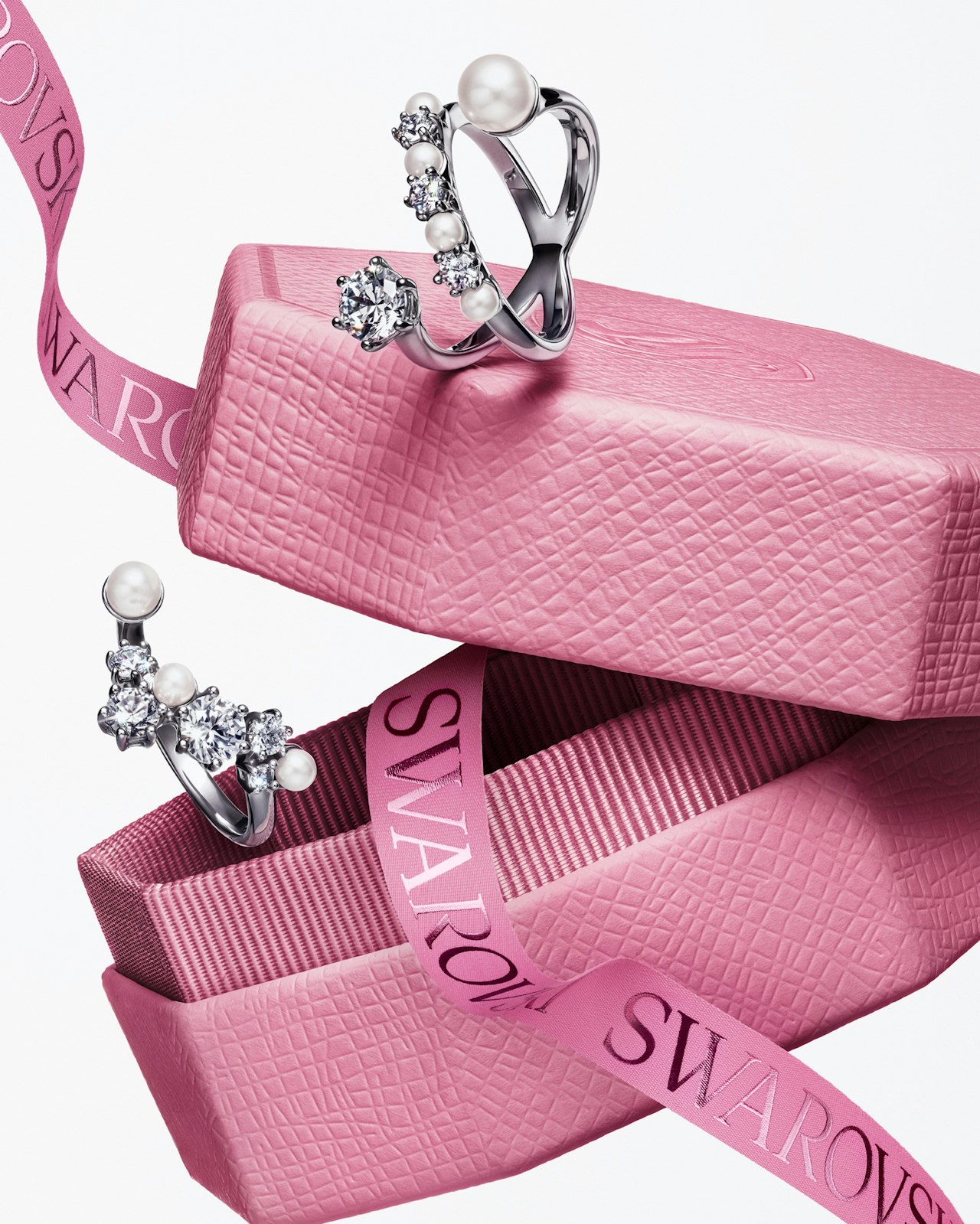 Two silver rings with pearls and gems on stacked pink boxes and a Swarovski ribbon.