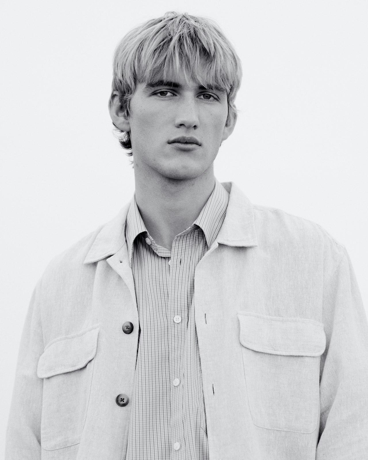 Young man with light hair wearing a buttoned shirt and light jacket, looking forward.