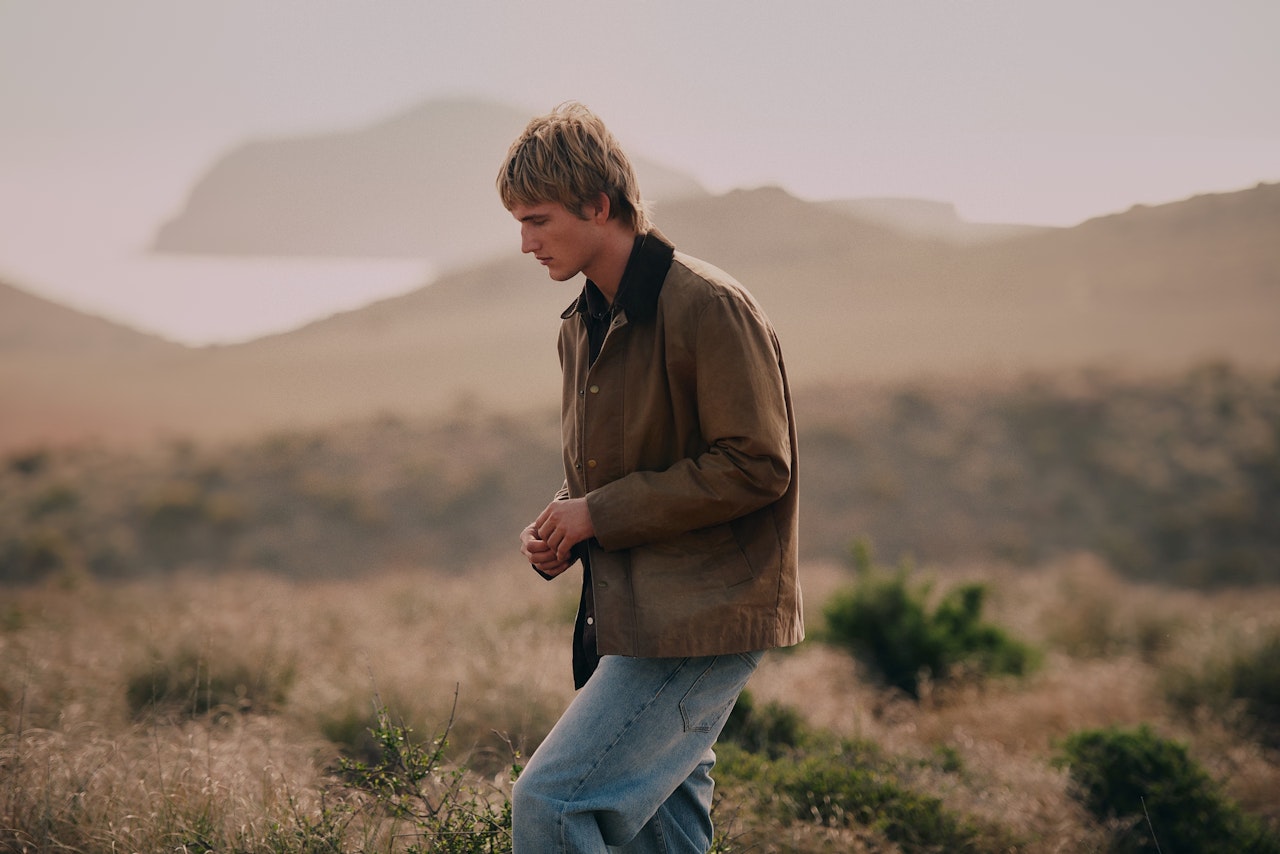 Person in a brown jacket walking through a grassy field with hills in the background.