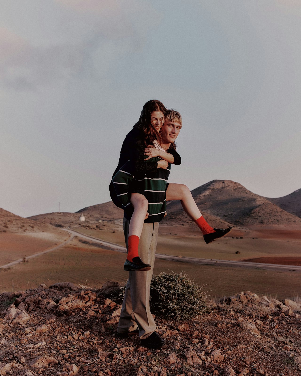 A person carries another on their back in a rocky, desert landscape at sunset.