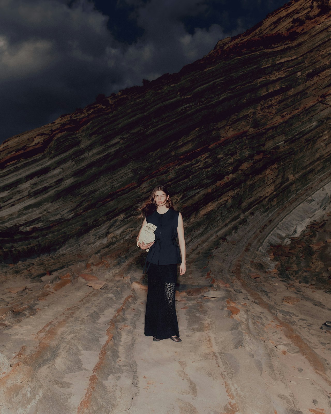 Woman in black outfit stands on rugged, sloped rock under a dark, cloudy sky.