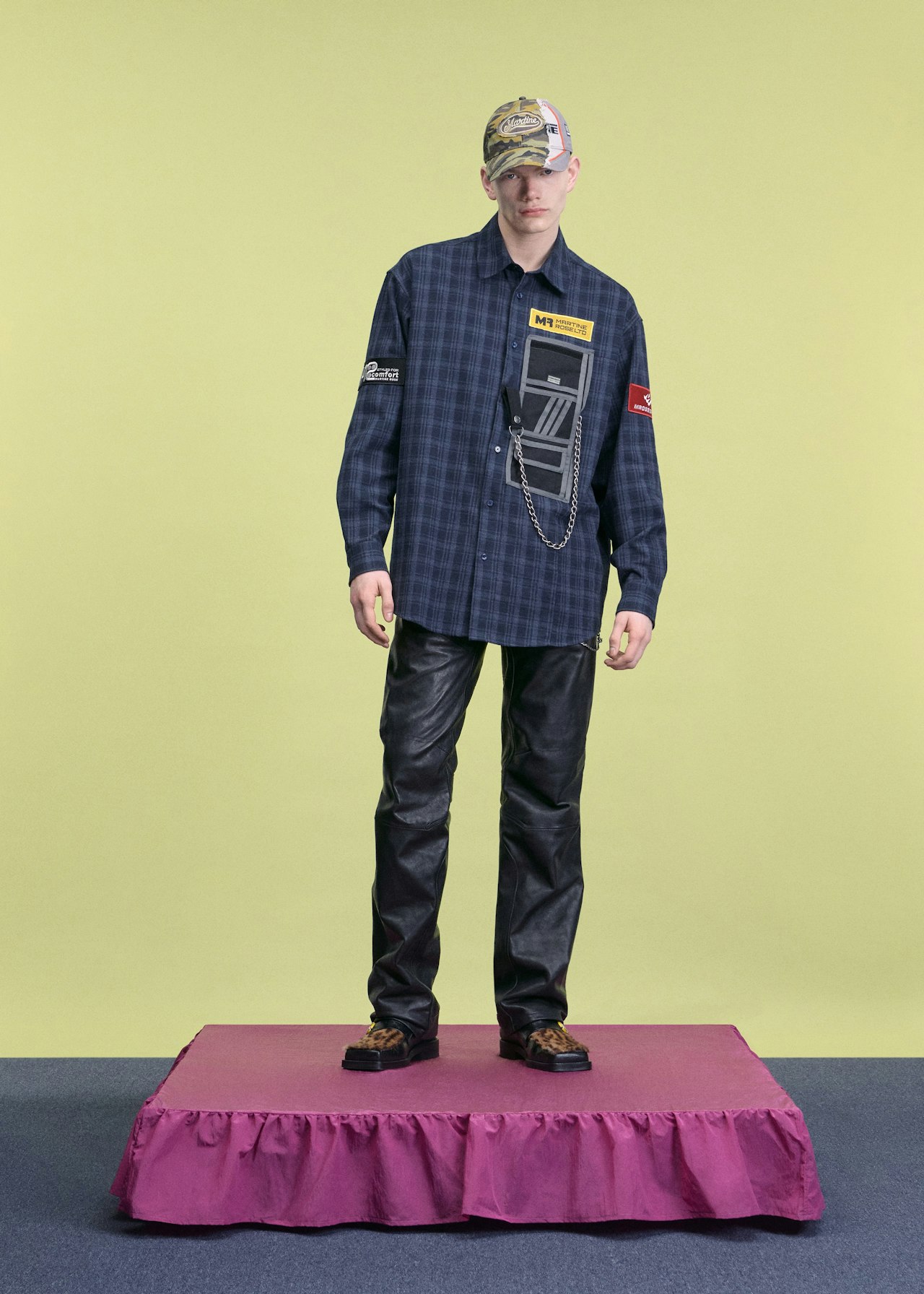 Person in a plaid shirt, leather pants, and cap stands on a small pink platform.