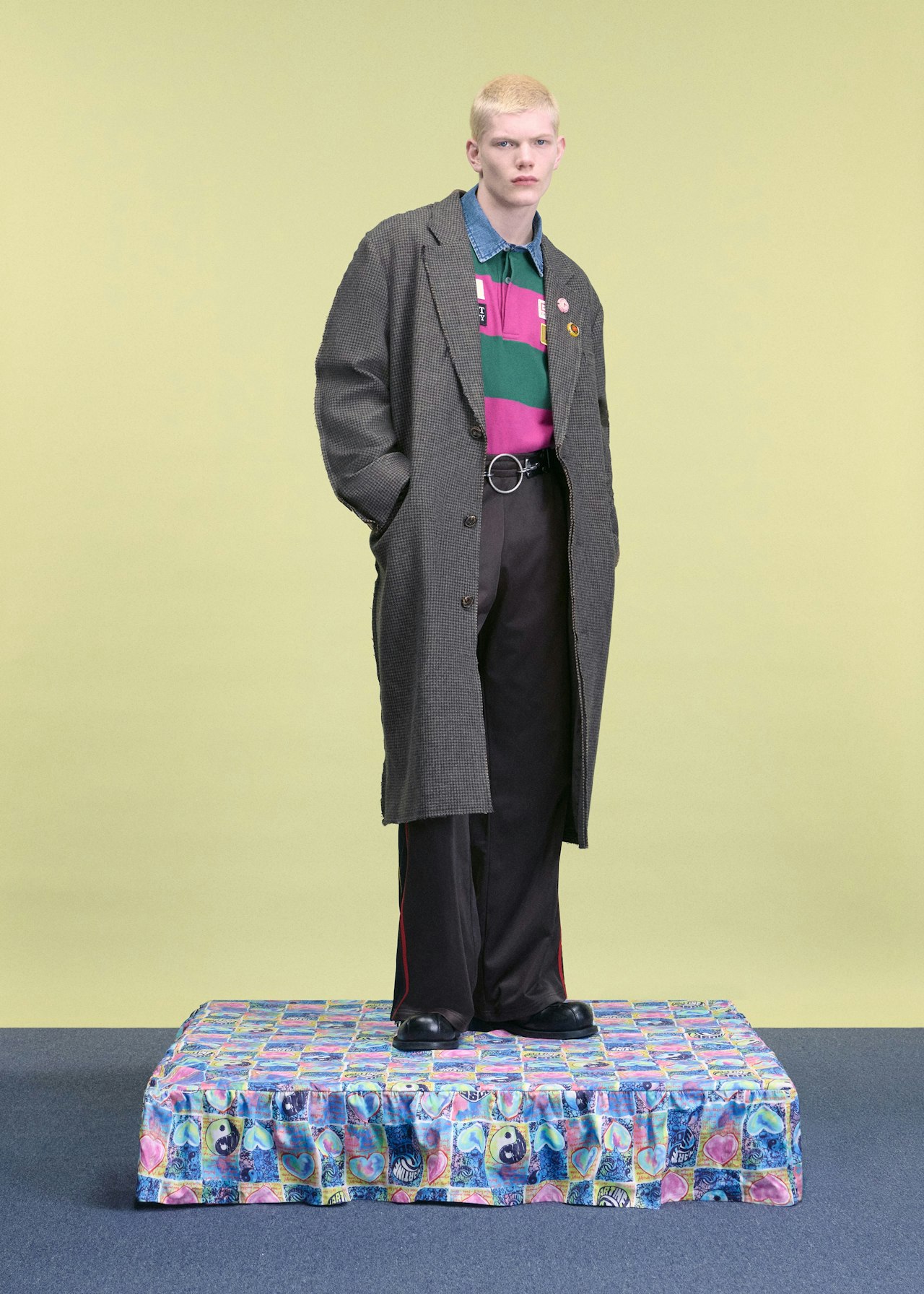 Person in a long coat standing on a colorful platform against a pale background.