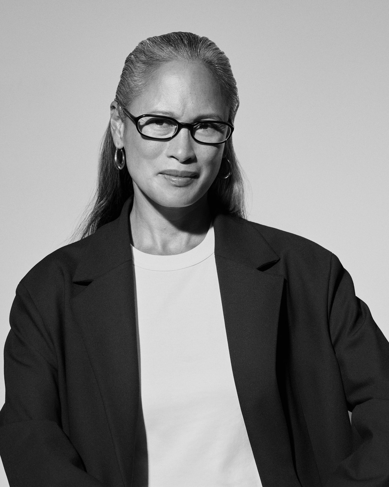 A woman with glasses and hoop earrings wearing a blazer looks at the camera.