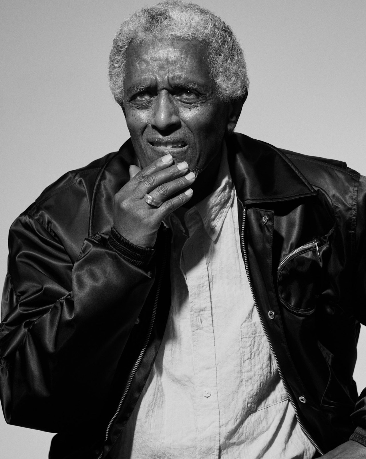 Elderly man in a jacket and shirt, touching his mouth, looking pensive.