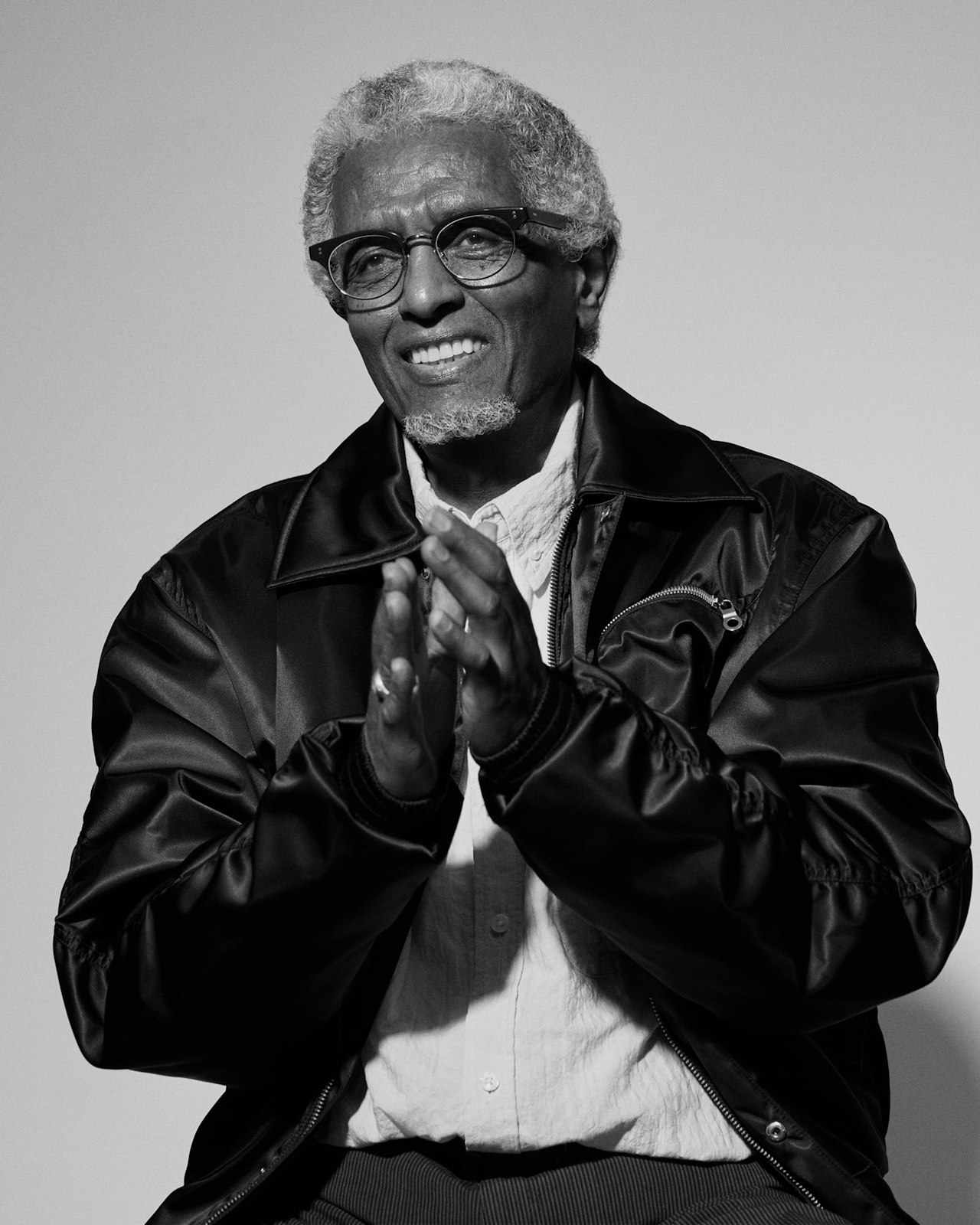 Older man with glasses smiles and claps, wearing a jacket and light shirt.