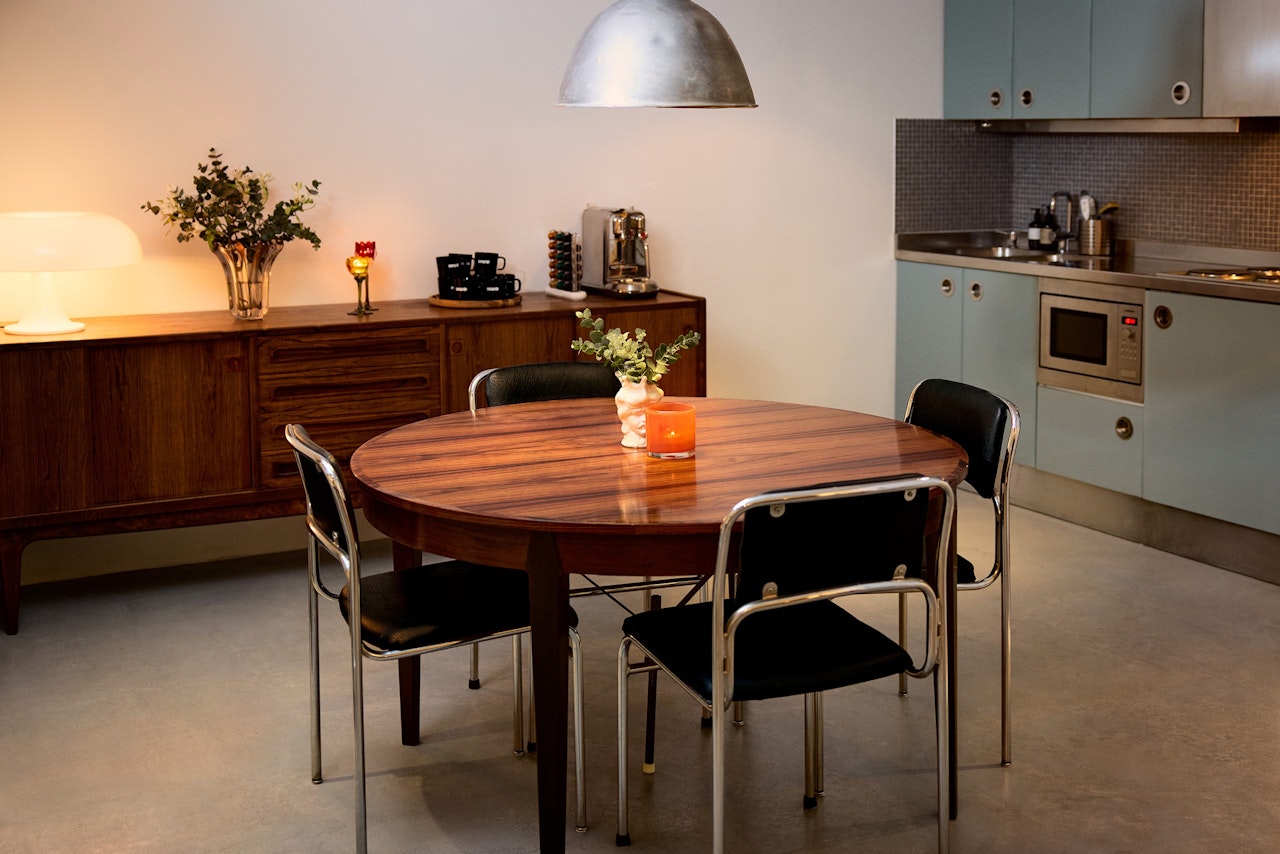 A modern kitchen with light blue cabinets, stainless steel appliances, and a round wooden dining table with four chairs; a sideboard with decor items is against the wall.