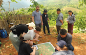 Lao and Swiss participants gather outdoors around a large printed map during a field activity in a rural, hilly landscape. Some are kneeling and marking the map, while others observe and discuss the area, surrounded by trees and mountain views.