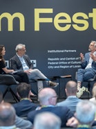 Three people sitting on stage during a fireside conversation, with an audience watching.