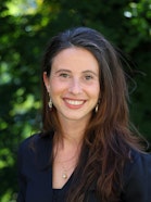 A person in a black shirt with a bright smile, hair pulled to the side.