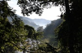 A rocky river runs through a forested valley in northeastern Madagascar, framed by dense tropical vegetation in the foreground and mist-covered hills rising in the distance.