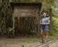 A young woman in a grey K'erenda Homet shirt stands smiling on a wooden walkway in front of the carved 'Refugio K'erenda Homet' sign with its bird motifs.