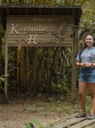 A young woman in a grey K'erenda Homet shirt stands smiling on a wooden walkway in front of the carved 'Refugio K'erenda Homet' sign with its bird motifs.