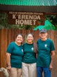 Two women and an older man in matching green K'erenda Homet shirts smile together beneath the wooden 'Área de Conservación Privada K'erenda Homet' entrance sign, a pale dog beside them