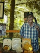 An older man in a plaid shirt and cap holds a machete over his shoulder beside a display of framed photographs, certificates, and conservation awards.