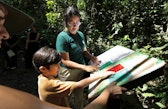 A woman in a green K'erenda Homet shirt and a young boy point at a colorful interpretive panel along a shaded forest trail, with other visitors nearby.