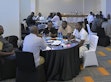 Several small groups seated at round tables in discussion in a workshop room; a participant in a Kenya jersey in the foreground.