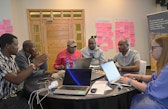 Sechs Workshop-Teilnehmende an einem runden Tisch mit Laptops, einer gestikuliert beim Sprechen; an der Wand dahinter rosa Haftnotizen und ein Banner der Wyss Academy.