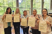 Five women stand together smiling and holding cloth bags printed with the Spanish phrase “Mujeres que están cambiando la minería.”