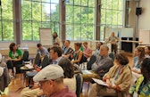 Conference participants sit in a bright room during a discussion session, with one person speaking into a microphone.
